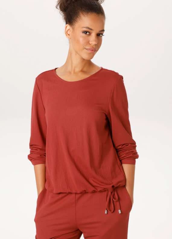 Frau in rotem Langarmshirt und passender Hose mit Händen in den Taschen.