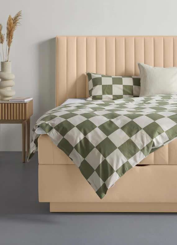 Bett mit beige gepolstertem Kopfteil, grüner und weißer Karobettwäsche, zwei Kissen, Nachttisch mit Vase und Pampasgras.