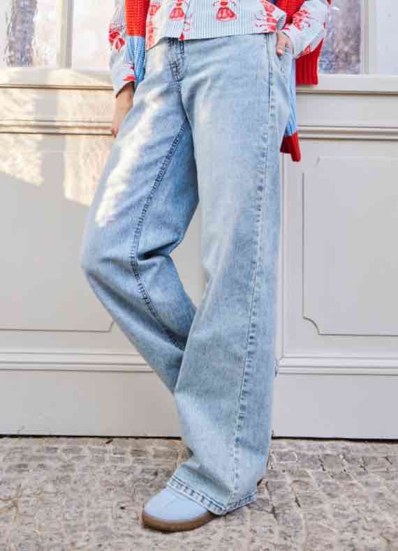 Person trägt weite, hellblaue Jeans und hellblaue Schuhe, steht vor einem hellen Hintergrund.