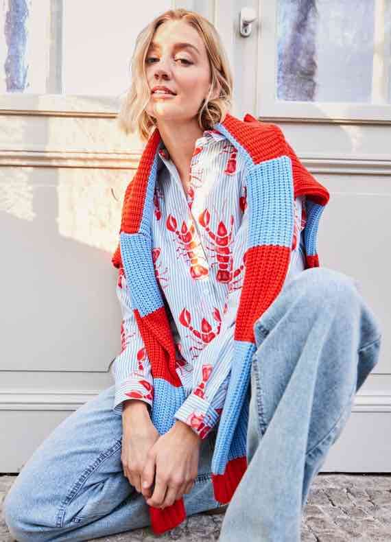 Person trägt Hemd mit Hummermuster und rot-blauen Pullover, sitzt auf dem Boden vor einer Tür.