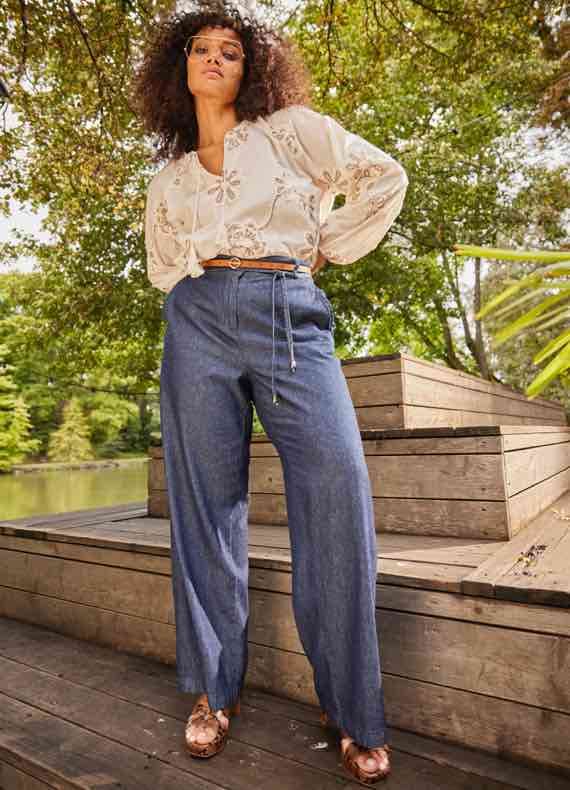 Frau in bestickter Bluse und blauen Hosen steht draußen auf Holzplattform, umgeben von grünen Bäumen.