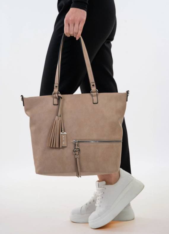 Person in schwarzen Hosen und weißen Sneakern hält beige Handtasche mit Fransen und Reißverschluss.