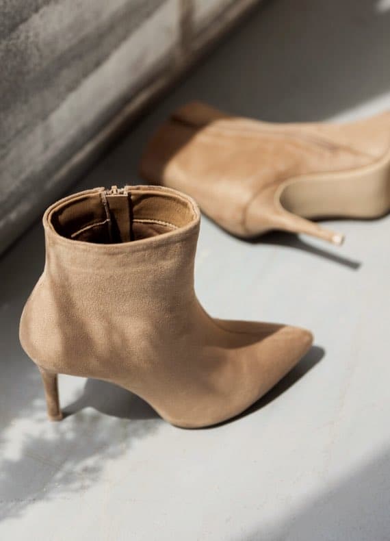 Beige Stiefeletten mit Absatz, eine liegend und die andere stehend, auf hellem Untergrund.