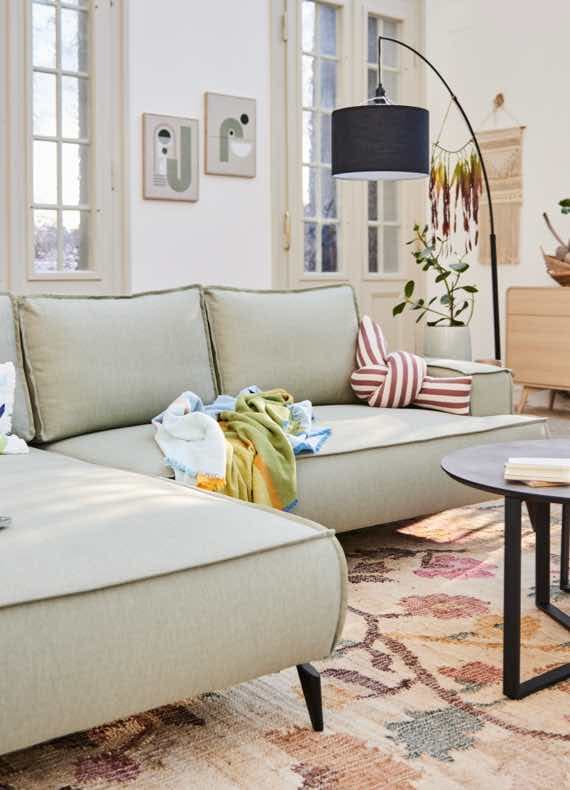 Gemütliches Wohnzimmer mit hellgrünem Sofa, bunten Kissen, Decken, Stehlampe und Wanddekorationen.