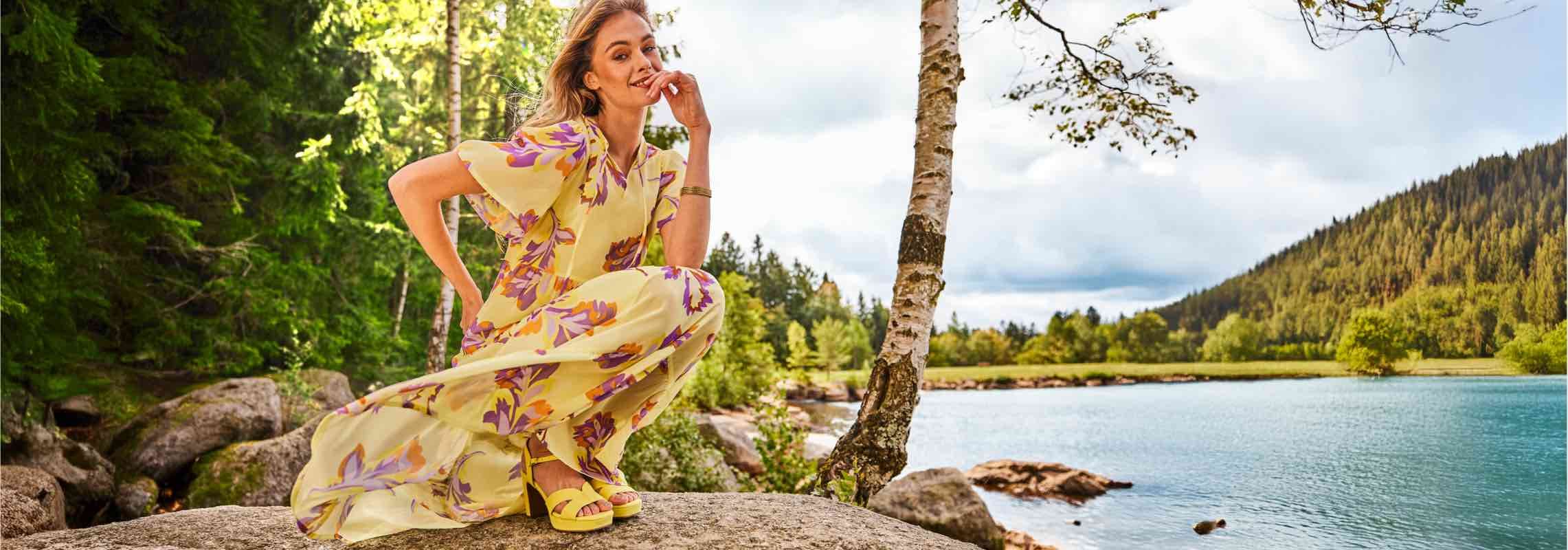 Frau in gelbem Kleid mit violettem Blumenmuster, kniet auf einem Felsen vor einem See und Wald.