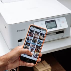 Hand hält Smartphone mit Fotogalerie vor weißem Canon-Drucker