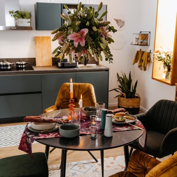 Küchentisch mit gedeckten Tellern, Gläsern, Kerze und Blumenarrangement. Moderne Küche im Hintergrund.