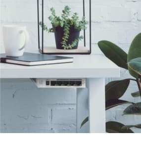 Schreibtisch mit Buch, Tasse, Pflanze und kleinem Netzwerkgerät darunter, an weißer Ziegelwand.