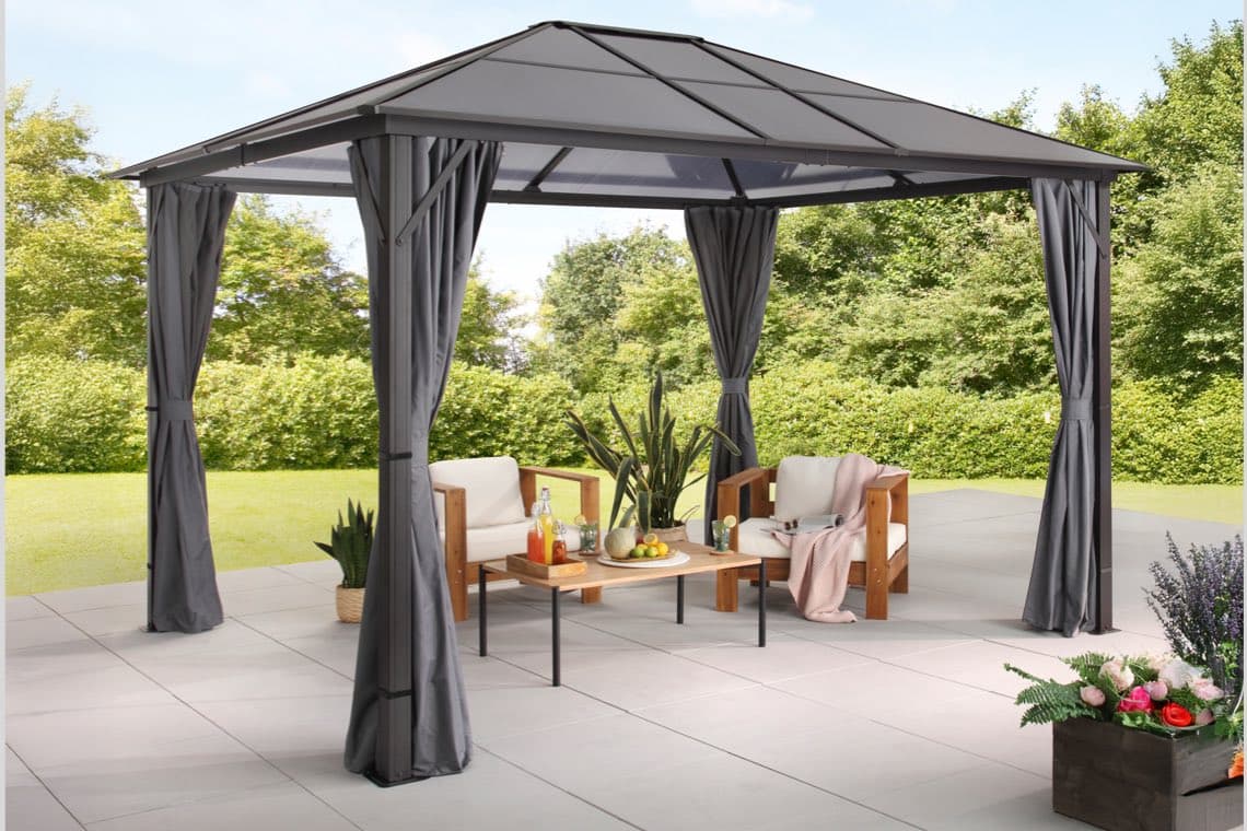 Gartenpavillon mit grauen Vorhängen, zwei Sesseln und Tisch mit Getränken auf Terrasse vor grüner Hecke