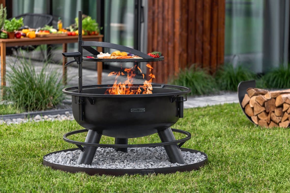 Runde Feuerschale im Garten, darüber Grillaufsatz mit Gemüse und Brot, im Hintergrund Holztisch mit Zutaten und Holzstapel