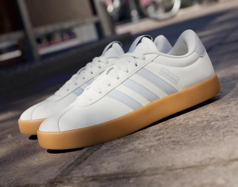 Adidas-Sneaker in Weiß mit hellbrauner Sohle und blauen Streifen auf einem Steinboden.