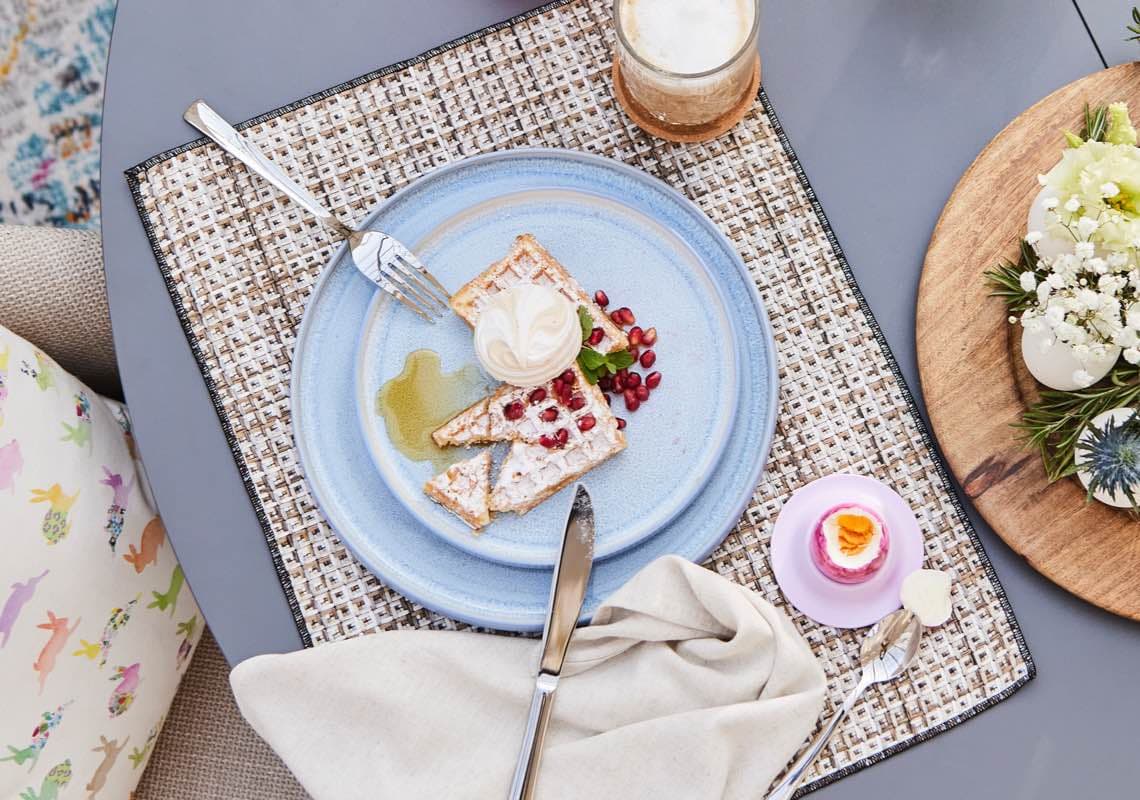 Waffeln mit Sahne und Granatapfelkernen auf blauem Teller, daneben Getränk und Dessert auf Tischmatte.