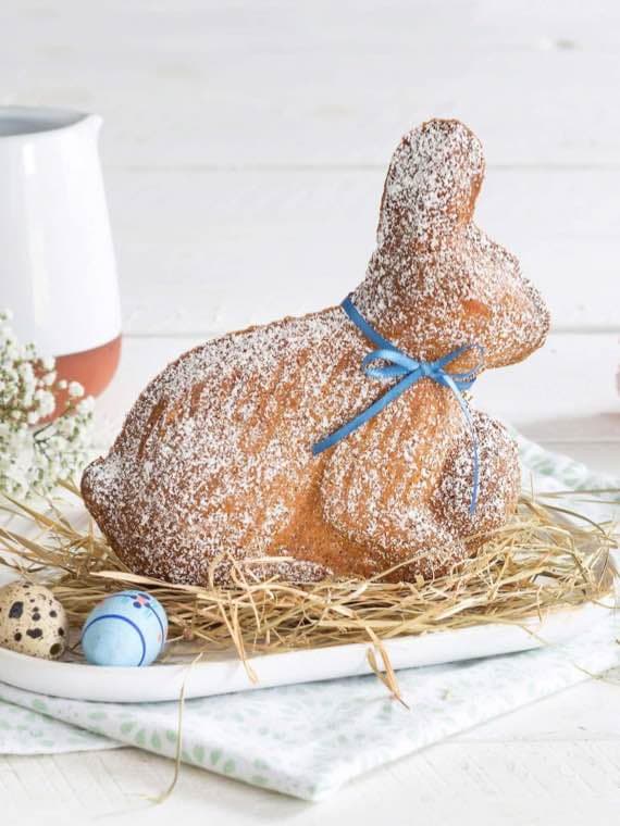 Gebäck in Form eines Hasen mit blauem Band, bestäubt mit Puderzucker, auf Heu, neben bemalten Ostereiern.