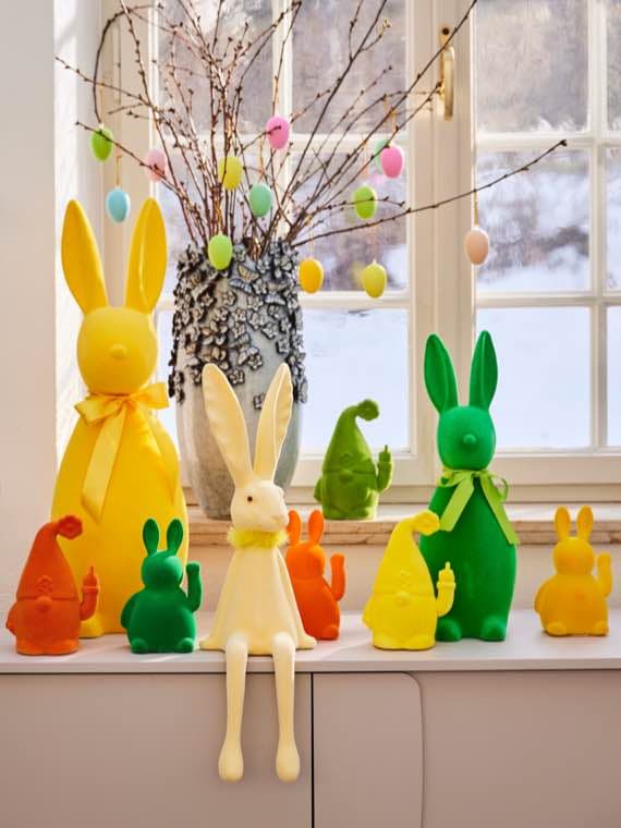 Bunte Osterdekoration mit Hasenfiguren vor einem Fenster. Vase mit Zweigen und bunten Eiern.