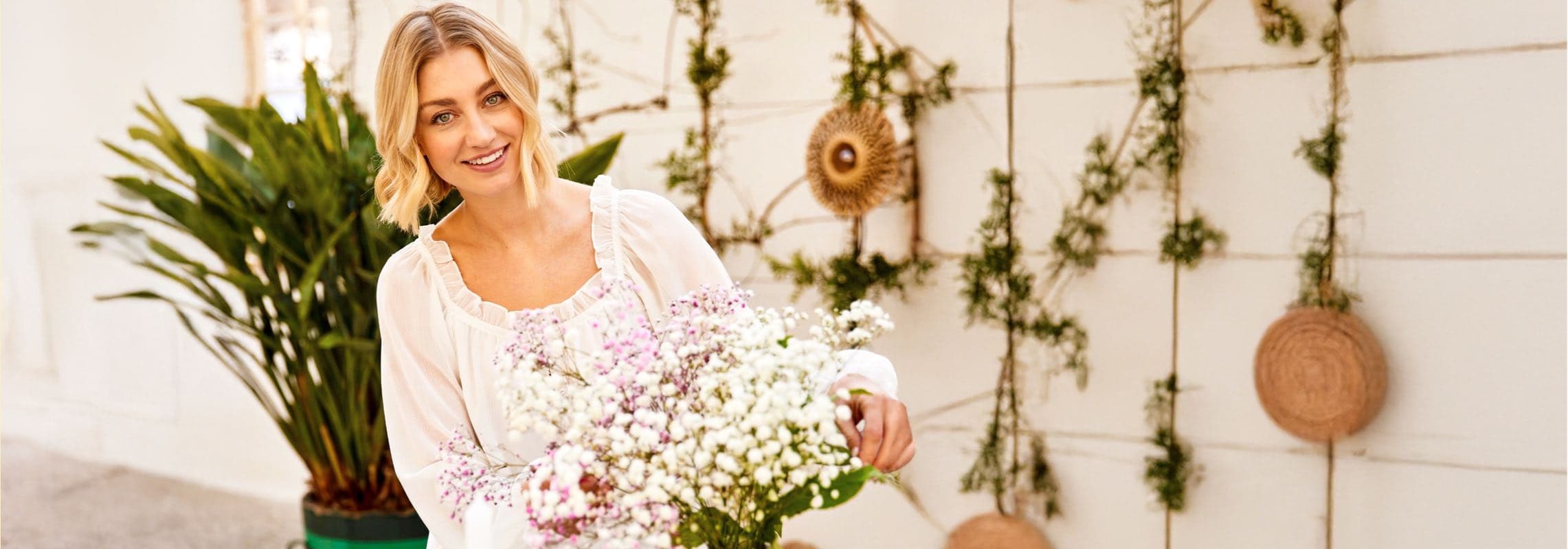 Frau in heller Bluse hält großen Strauß weißer und rosa Blumen vor dekorativer Wand mit Ranken und Körben