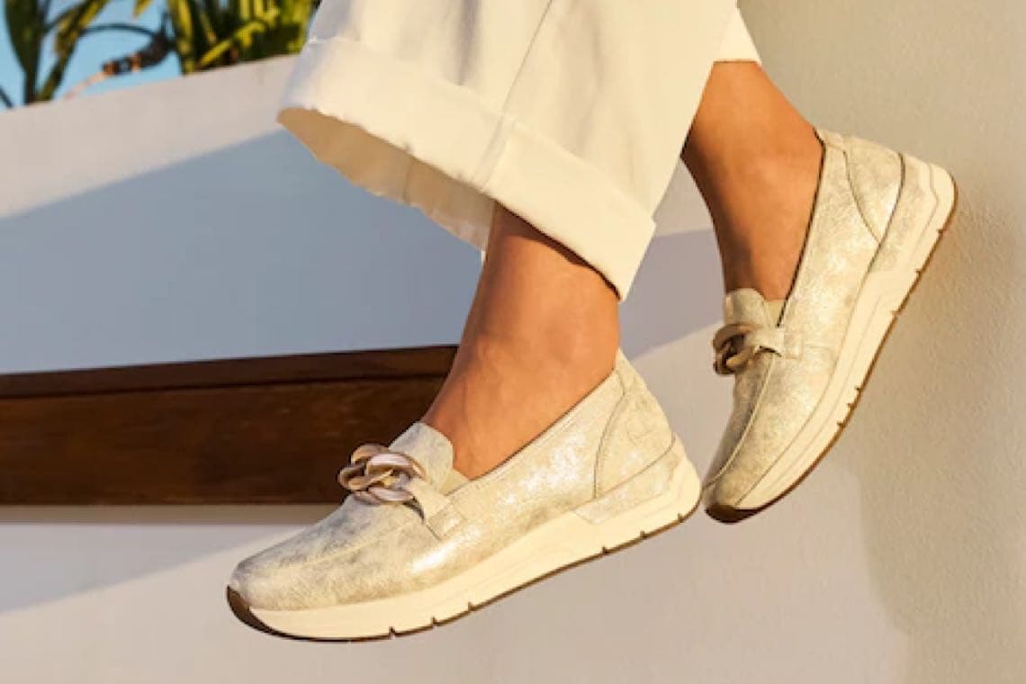 Beige Slipper mit Kette am Spann an Füßen in hochgekrempelter, heller Hose vor sonniger Hauswand