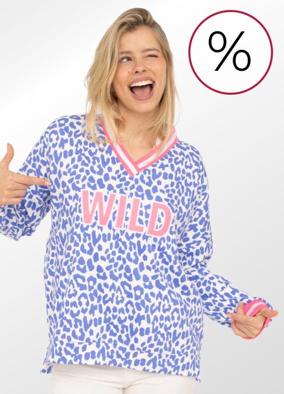 Frau im gemusterten Pullover mit Aufdruck WILD zeigt mit beiden Händen auf die Schrift