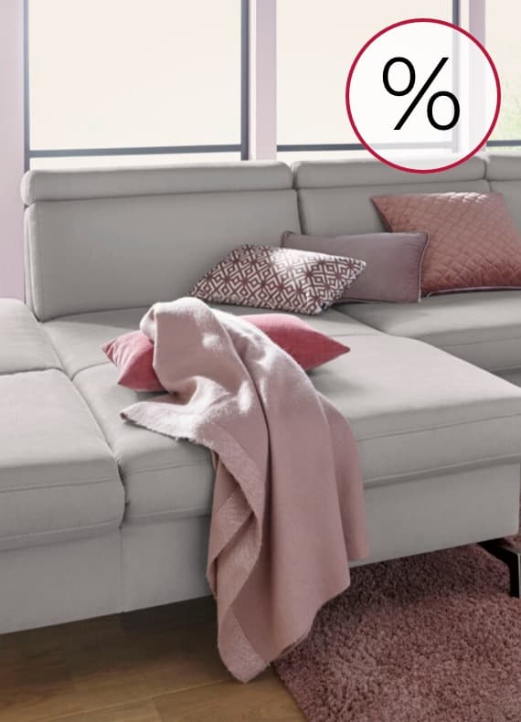 Prozentzeichen für Rabatt – großes Ecksofa mit rosa Decke und Kissen vor hellen Fenstern