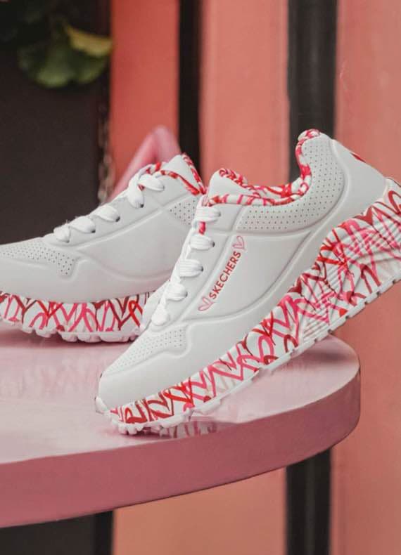 Skechers Logo auf weißen Sneakers mit pink-rotem Muster an Sohle und Futter, präsentiert auf rundem Tisch vor rosa Wand