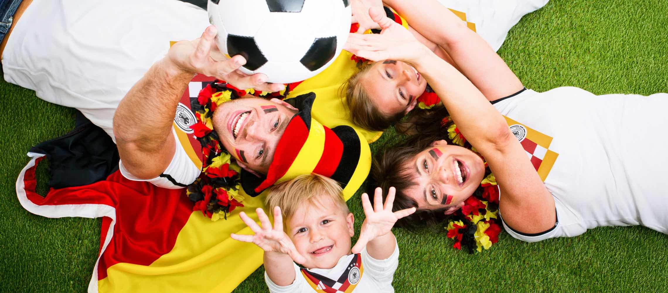 Menschen liegen auf Gras und halten einen Fußball. Sie tragen Deutschland-Fanartikel und lächeln.
