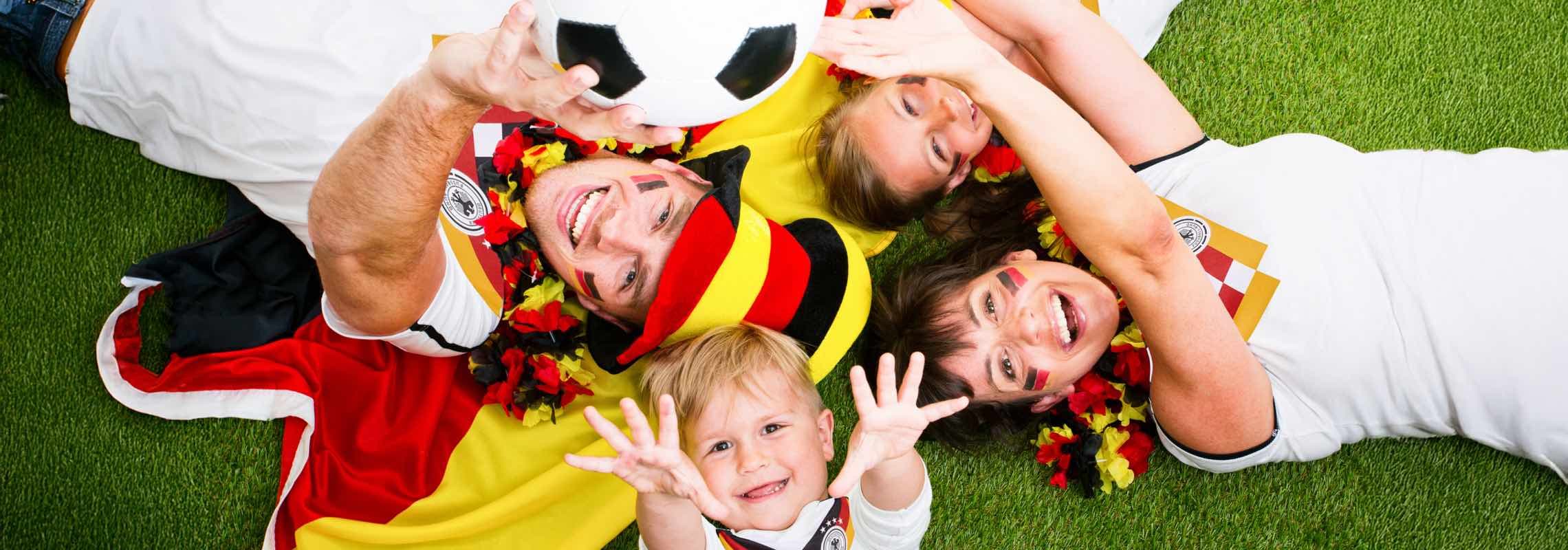 Familie in Deutschland-Trikots liegt auf Rasen, hält Fußball in die Kamera und trägt Deutschland-Schals und -Hüte