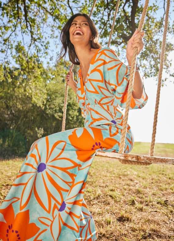 Frau auf Schaukel im Garten trägt langes Kleid mit großem orange-lila Blumenmuster auf hellblauem Hintergrund