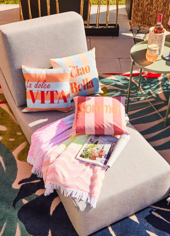 Kissen mit Aufdruck „La dolce Vita“, „Ciao Bella“ und „Sommer“ auf Liege mit Decke, daneben Tisch mit Getränk