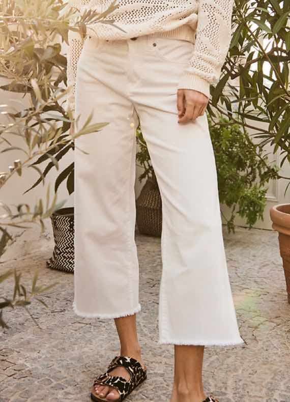 Frau trägt weite weiße Cropped-Hose mit ausgefranstem Saum, dazu Strickpullover, steht zwischen Pflanzen draußen