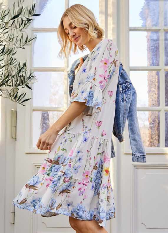 Frau im geblümten, knieumspielenden Kleid schwingt den Rock, Jeansjacke locker über der Schulter, helle Fenster im Hintergrund