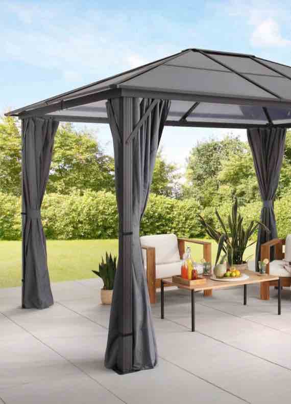 Gartenpavillon mit grauen Vorhängen auf Terrasse, darunter Sitzgruppe mit Tisch, Getränken und Obst im Grünen