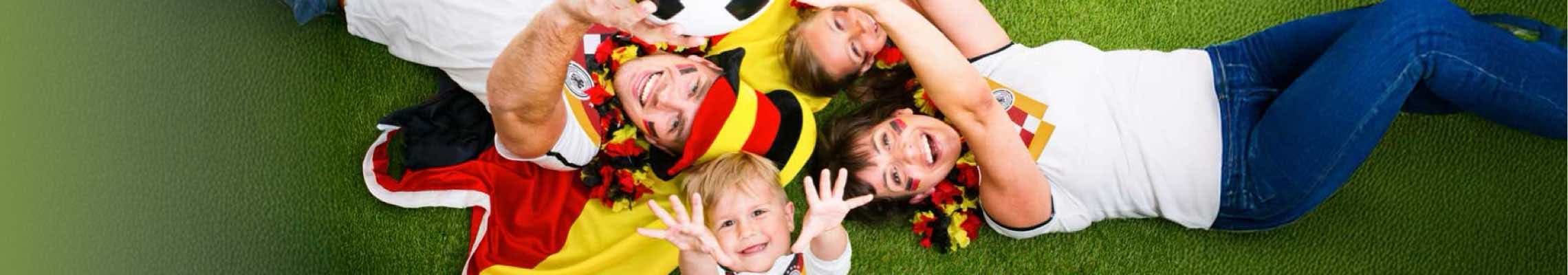 Familie in Fan-Kleidung liegt mit Deutschlandflagge und Fußball auf dem Rasen, Kind streckt die Arme nach oben