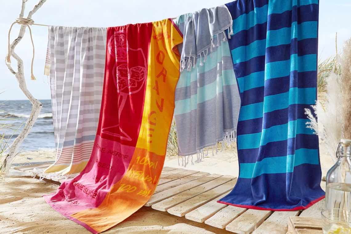 Bunte Strandtücher mit Streifen und Cocktailmotiv hängen auf Leine über Holzsteg am Meer