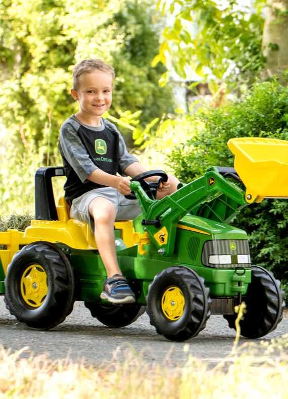 Junge fährt draußen auf grün-gelbem Kindertraktor mit Frontlader und Anhänger auf einem Weg