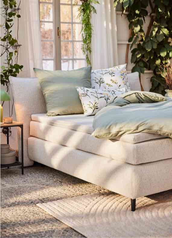 Helles Bett mit grüner Bettdecke und gemusterten Kissen vor Fensterfront, umgeben von vielen Zimmerpflanzen