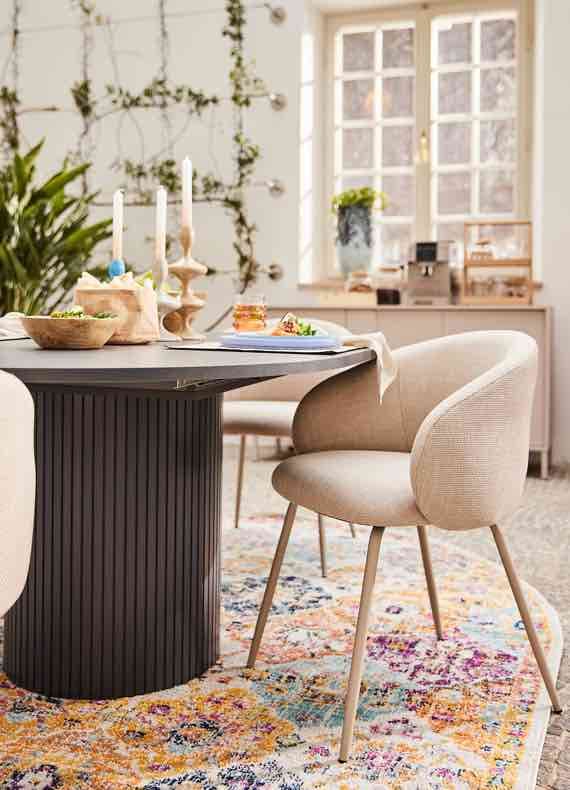 Runder Esstisch mit Gedeck und Kerzen, beigefarbener Stuhl auf buntem Teppich in hellem Esszimmer mit Fenster im Hintergrund