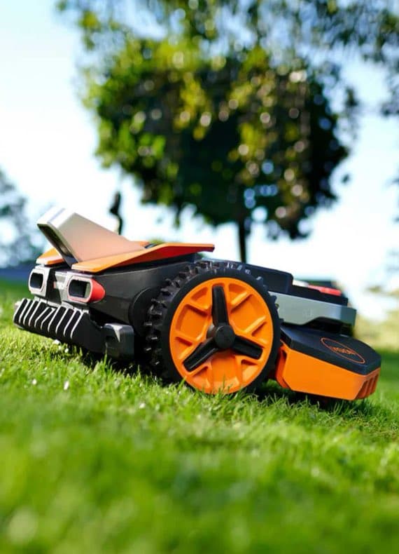 Oranger Mähroboter fährt über frisch geschnittenen Rasen im Garten, Bäume verschwimmen im Hintergrund
