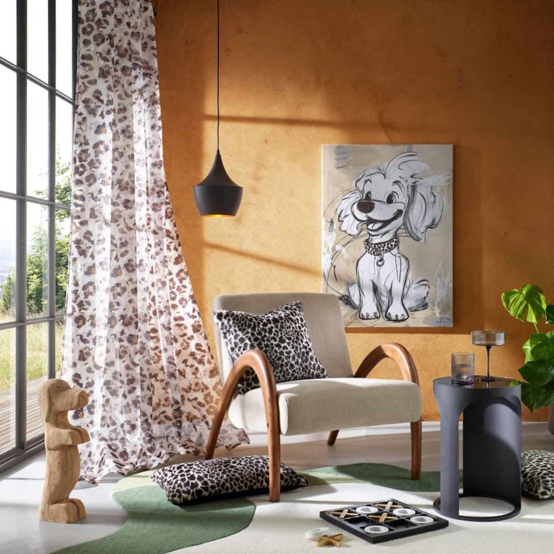 Wohnzimmer mit Sessel, Leopardenmuster-Gardine, Kissen, Hundebild an der Wand und kleinem Beistelltisch mit Deko