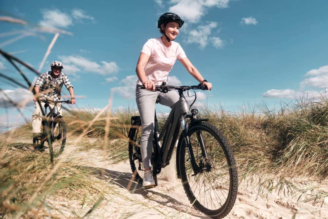 Zwei Personen mit Fahrradhelmen fahren mit E-Bikes auf sandigem Dünenweg unter blauem Himmel