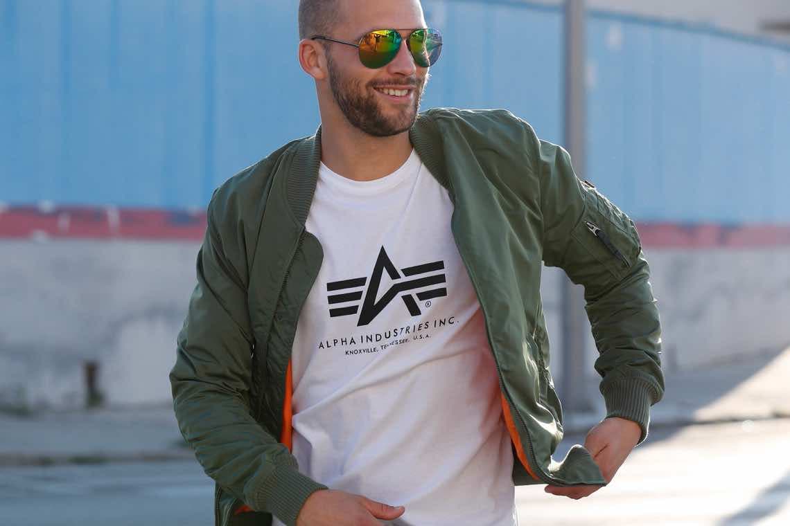 ALPHA INDUSTRIES INC. Logo – Person im weißen Logo-T-Shirt mit geöffnetem grünen Blouson auf der Straße