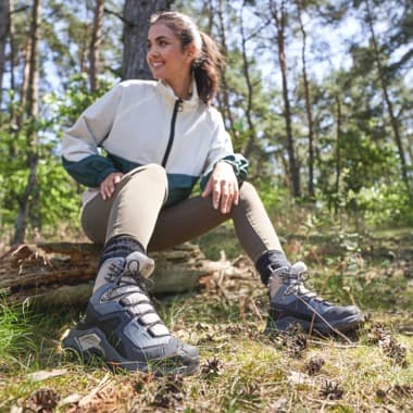 Frau sitzt im Wald auf einem Baumstamm und präsentiert graue Wanderschuhe auf grasigem Waldboden