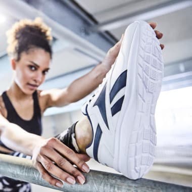 Person dehnt sich an einer Stange und streckt weißen Sportschuh mit dunklen Streifen in die Kamera