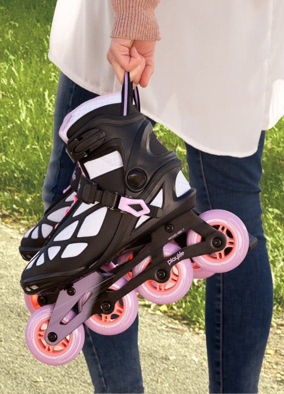 Person hält ein Paar Inline-Skates mit rosa Rollen in der Hand, steht in Jeans auf einem Weg neben einer Wiese