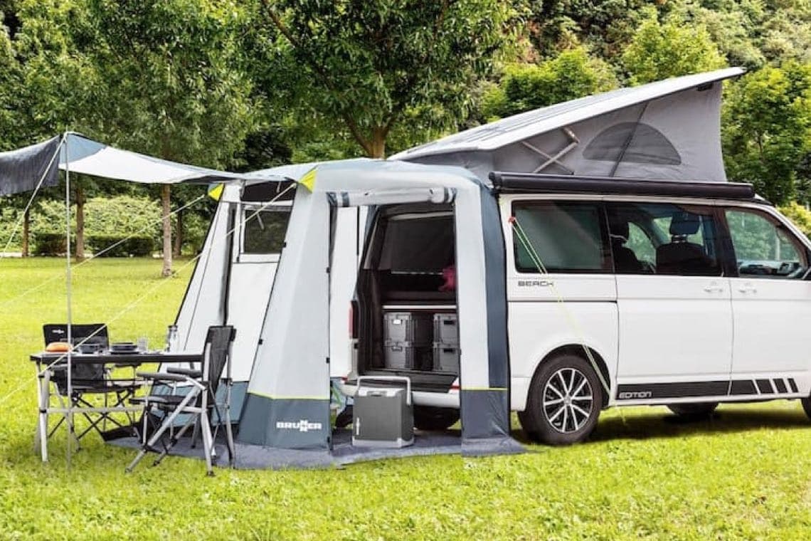 Campingbus mit aufgebautem Vorzelt, Markise und Campingtisch mit zwei Stühlen auf einer grünen Wiese