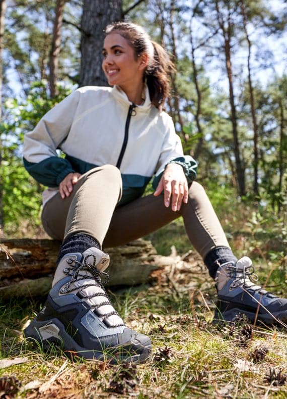 Frau mit Outdoorjacke sitzt im Wald auf einem Baumstamm und zeigt graue Wanderstiefel mit Schnürung
