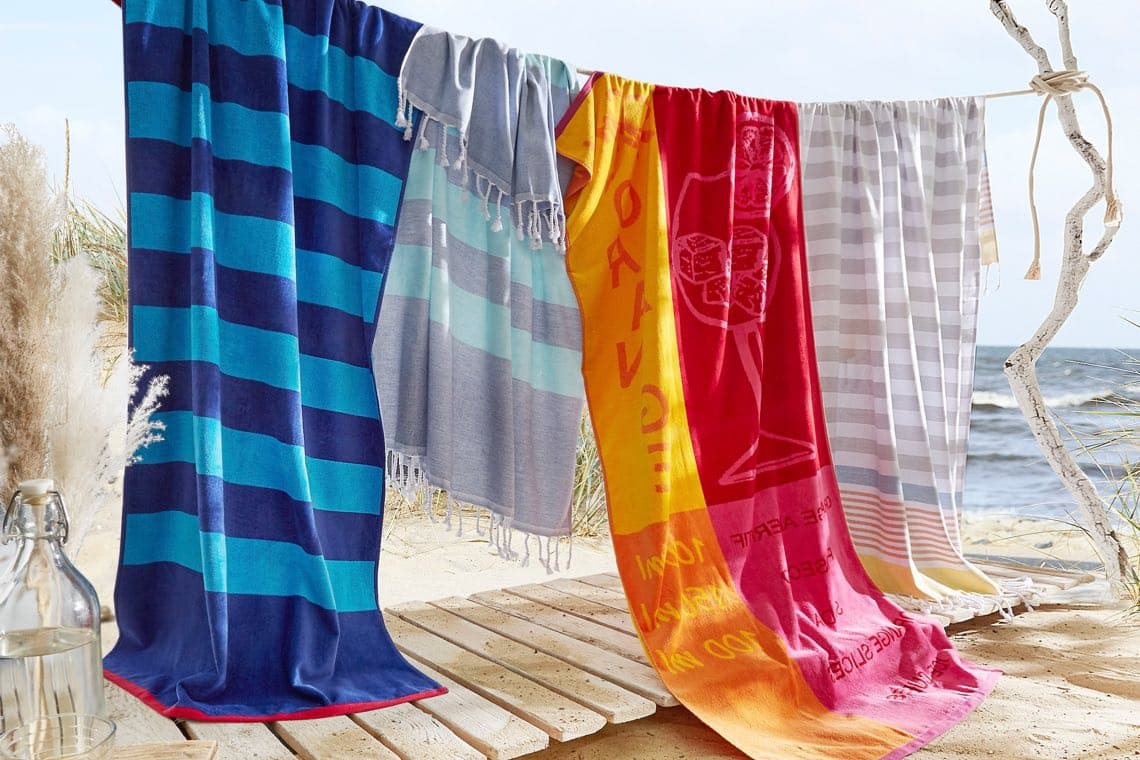 Bunte Strandtücher mit Streifen und Motiv hängen auf Leine am Holzsteg vor Meer und Sandstrand