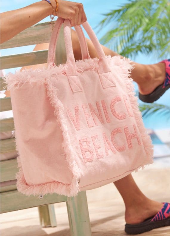 VENICE BEACH – große rosa Strandtasche mit Fransen, von einer Person auf einer Bank am Strand gehalten