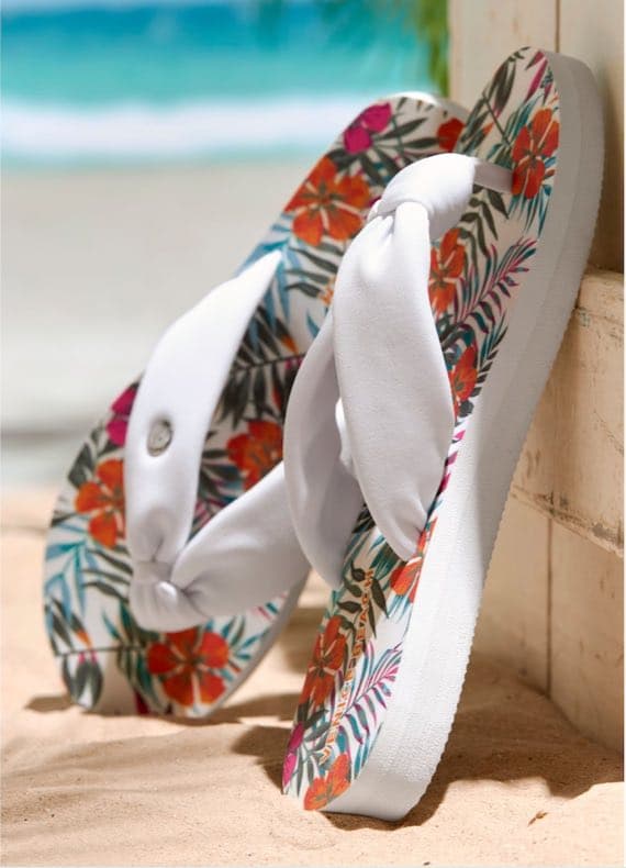Zehentrenner mit buntem Blumenmuster im Sand, im Hintergrund verschwommener Strand mit Meer
