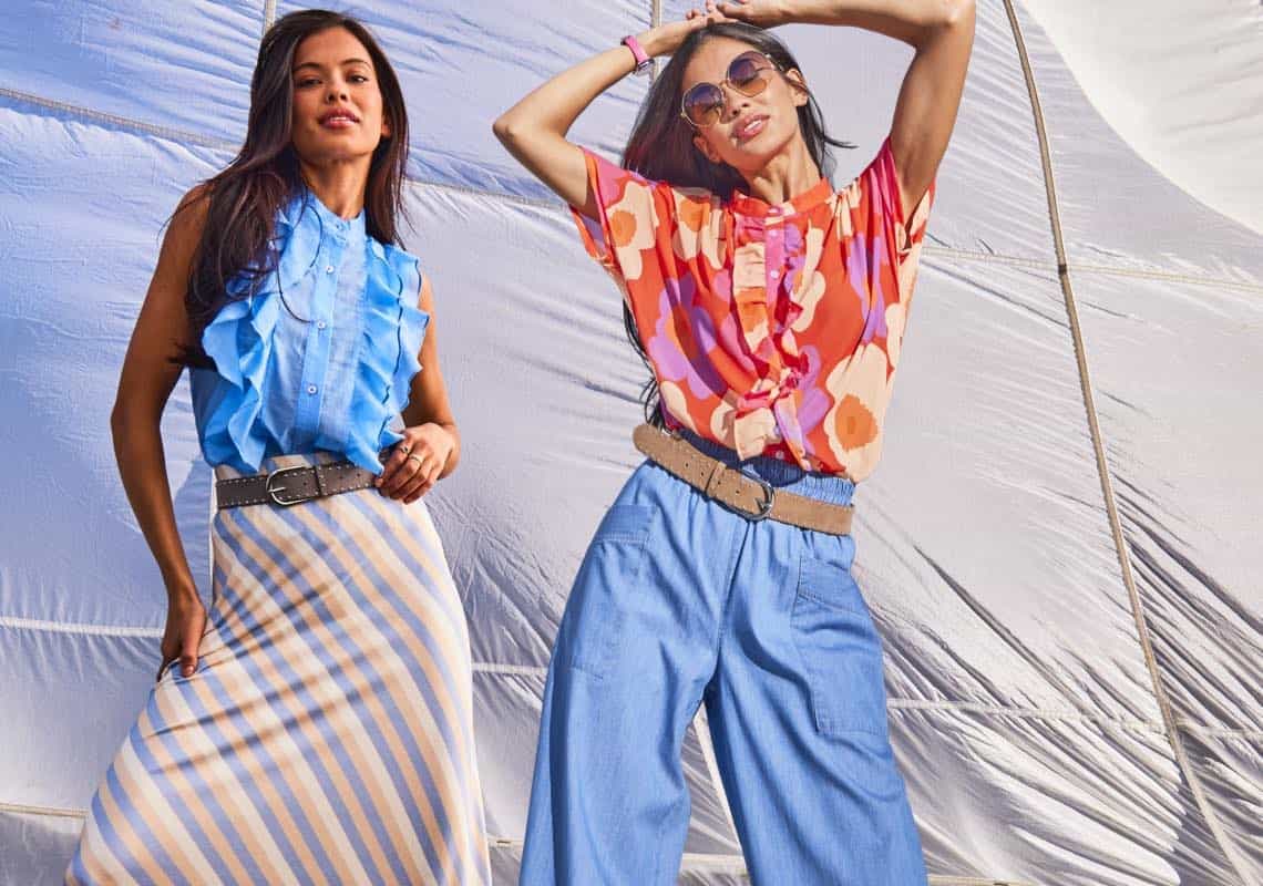 Zwei Frauen posieren vor hellem Stoffzelt in bunten Sommeroutfits mit Rüschenoberteilen, weitem Rock und lockeren Hosen