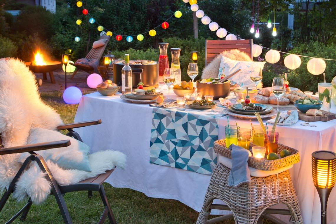 Sommerliche Gartentafel mit Getränken, Speisen und Lichterketten, daneben Feuerstelle und gemütliche Liegestühle