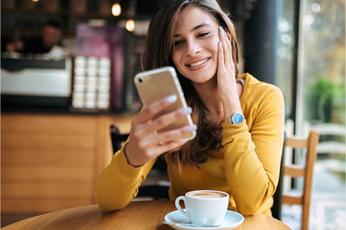Frau im gelben Oberteil sitzt im Café, schaut auf ihr Smartphone, vor ihr eine Tasse Kaffee auf dem Holztisch